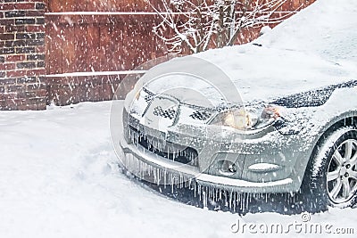 Closeup of front of silver car in snowstorm with lights on but covered in snow and icicles Stock Photo