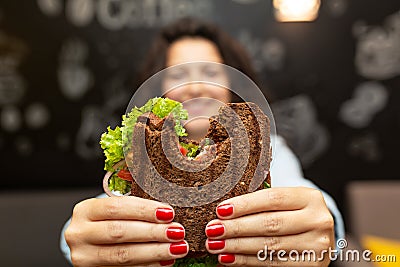 Closeup funny blurred protrait of young woman hold bitten sandwich by her two hands. Sandwich in focus. dark background Stock Photo