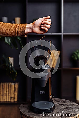 Closeup of the hand of a girl who pours out coffee beans into a black stylish coffee grinder. Coffee beans slowly fall into the Stock Photo