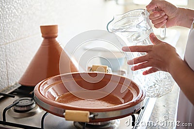 Closeup housewife beginning cooking meal in tajine clay pot, pouring some water inside a clay dish standing on a stove Stock Photo