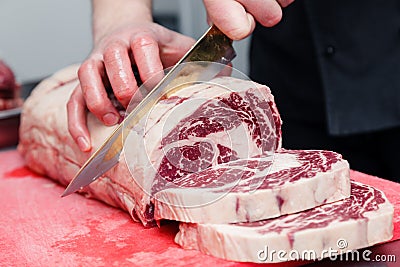 Closeup macro cook`s hand cut ribeye marbled beef steak with knife on red plastic cutting board on metal table in restaurant Stock Photo