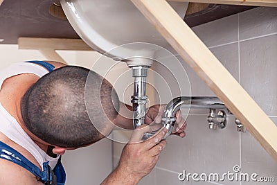 Closeup male plumber worker in blue denim uniform, overalls, fixing sink in bathroom with tile wall. Professional plumbing repair Editorial Stock Photo