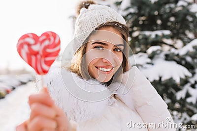 Closeup portrait amazing joyful smiling girl in sunny winter morning with pink lollypop on street. Attractive young Stock Photo