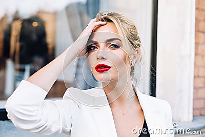 Closeup portrait attractive woman with red lips on street . She wears white jacket, touching hair, looking to camera. Stock Photo