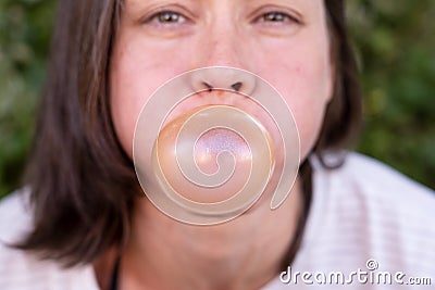 Closeup portrait of funny women, carefully inflates bubble gum, with narrowed eyes and round cheeks, outdoors. Stock Photo