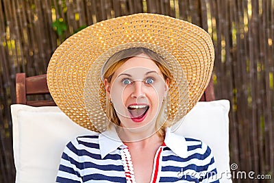 Closeup portrait of happy laughing unexpectedly young adult woman in hat and dress sitting on cozy deckchair and looking at camera Stock Photo