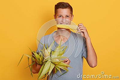 Closeup portrait of happy young student guy biting maize and holding other corns in hands, stands against yellow wall, attractive Stock Photo