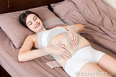Closeup pregnant woman dresses bandage on belly at home on bed. Orthopedic abdominal support belt Stock Photo