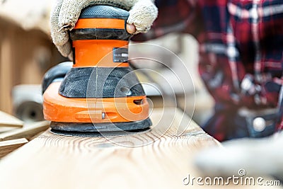 Closeup professional carpenter hand grinding raw wood plank with orbital sander electric machine in carpentry diy workshop. Detail Stock Photo