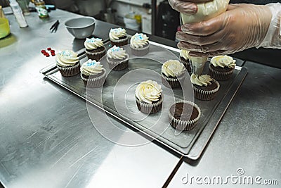 Closeup shot of person adding vanilla buttercream frosting with pastry bag onto chocolate muffins placed on stainless Stock Photo
