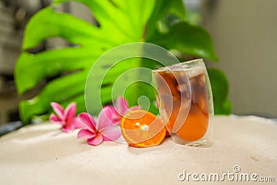 Closeup shot of a transparent glass full of orange juice next to a halved orange pink plumerias Stock Photo