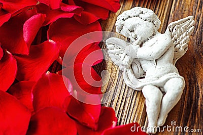 Closeup view of beautiful cupid with the trumpet, angel decorative figurine near red rose petals on wooden background. Stock Photo