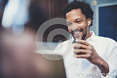 Closeup view of Smiling african man using smartphone to listen to music while sitting on the bench at sunny street Stock Photo
