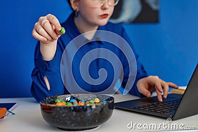 Closeup of young woman enjoying candy snack while using laptop in vibrant office Stock Photo