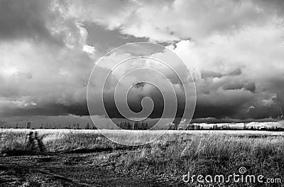 Cloudy sky before a thunderstorm, a field with a road going into the distance. Residential buildings near the horizon. Black and Stock Photo