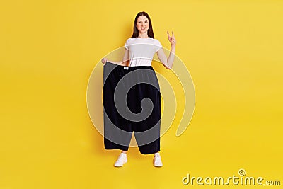 Cocky European woman with dark hair, wearing white t shirt and old too big pants, loosing weight, showing rock sing, looking at Stock Photo