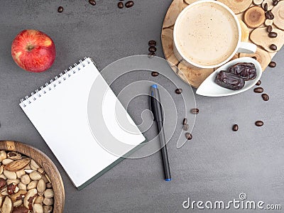 Coffee break in the workplace. White cup of coffee, apple, dates, mix of nuts in wooden bowl, notepad, pen on grey Stock Photo
