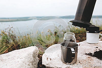 Coffee drops from aeropress on metal mug on cliff at lake, brewing alternative coffee at camping. Making hot drink at picnic Stock Photo