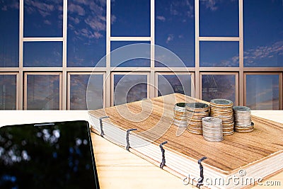 Coins stack on handmade book with tablet on wood table with building reflect sky background. Stock Photo