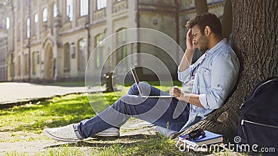 College student sitting under tree with laptop looking upset disappointing news Stock Photo