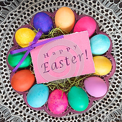 Colorful Easter Eggs on a Plate in center with Pink Happy Easter Name Tag Card on Vintage Lace Tablecloth. A Square crop with goo Stock Photo