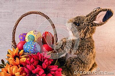 Colorful painted Easter eggs lie in a basket with flowers, on a wooden background, with a fluffy Easter bunny. Stock Photo