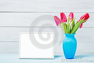 Colorful red spring tulip flowers in nice blue vase and blank photo frame on light wooden background as greeting card. Mothersday Stock Photo