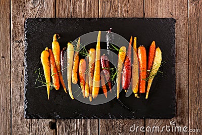 Colorful roasted rainbow carrots arranged in a row on a slate serving platter against a rustic wood background Stock Photo