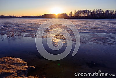 Colorful sunset with sun beams on the shore of a lake in early spring. Reeds and trees in the distance. The surface half-covered Stock Photo