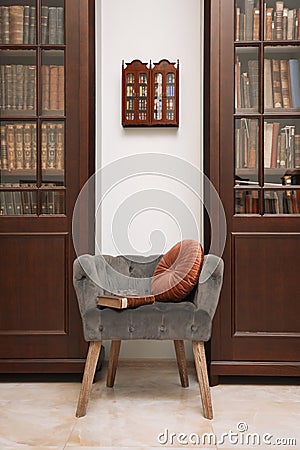 Comfortable armchair with pillow and book between wooden bookcases in library Stock Photo