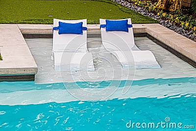 Comfy white loungers in shallow end of refreshing blue swimming pool in yard with flower trim around the edge Stock Photo