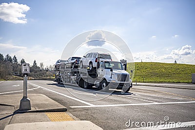 Compact white big rig car hauler semi truck transporting cars on the two level module semi trailer standing on the crossroad Stock Photo