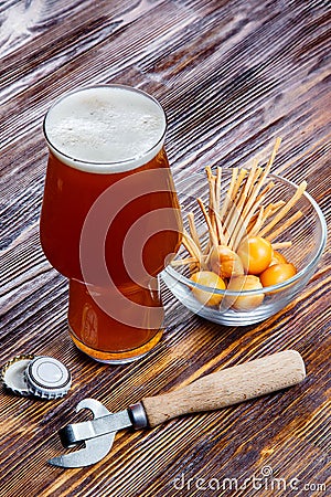 Composition of a glass of beer with foam on a rustic wooden table next to a bowl of salty snacks and a bottle opener Stock Photo