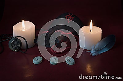 A composition of lighted candles, a bag of runes, an amulet with the image of Yggdrasil and a blue feather on a red background. Stock Photo