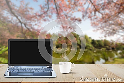 Computer laptop with black screen and hot coffee cup on wooden table top on blurred lake and cherry blossom tree background Stock Photo