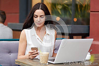 Concentrated beautiful female reads attentively information on web page via cell phone, sits in front of opened laptop computer, s Stock Photo