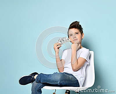 Concentrated schoolboy sits on chair holding dollars and touching chin with hand thinking how to earn more. Stock Photo