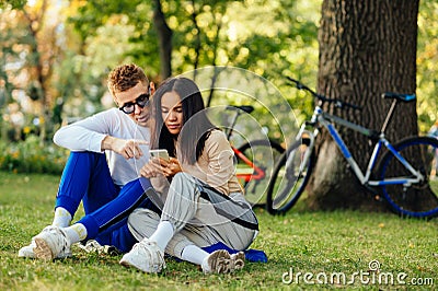 Concentrated couple is sitting on a grass in the park and using smartphone. A man is pointing a finger and showing something on Stock Photo