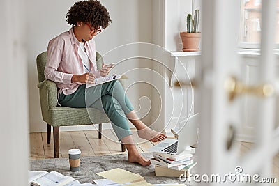 Concentrated dark skinned owner of business reads information from network, analyzes some strategies, sits on Stock Photo
