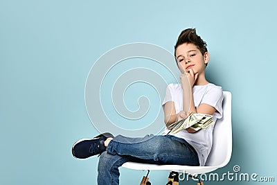 Concentrated schoolboy sits on chair holding dollars and touching chin with hand thinking how to earn more. Portrait isolated on Stock Photo