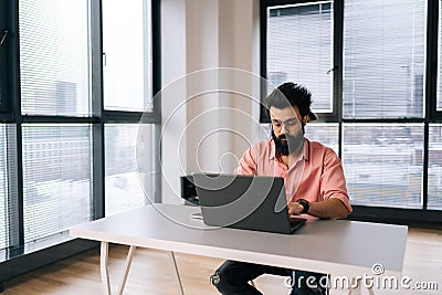 Concentrated Indian programmer male in eyeglasses working on laptop sitting at desk in light coworking office room by Stock Photo
