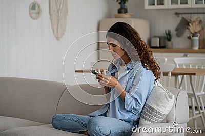 Concentrated introverted woman with smartphone sit on sofa with legs tucked under in living room Stock Photo