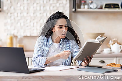 Concentrated lady sitting in kitchen, using laptop and reading book, studying remotely from home Stock Photo