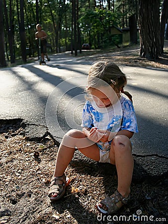 A concentrated little girl with a scratched knee sits on the asphalt in the summer Stock Photo