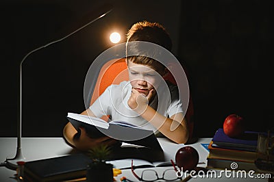 Concentrated schoolboy reading book at table with books, plant, lamp, colour pencils, apple, and textbook Stock Photo