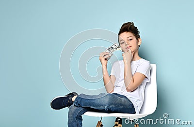 Concentrated schoolboy sits on chair holding dollars and touching chin with hand thinking how to earn more. Stock Photo