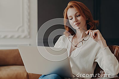 Concentrated serious redhead woman makes remote job indoors, sits in comfortable armchair, holds transparent glasses, reads Stock Photo