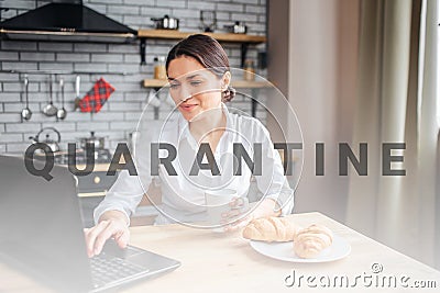Concentrated woman sit at table in kitchen and work. She typing on laptop keyboard. Woman hold hand on white cup Stock Photo