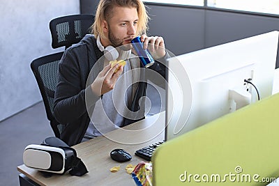 Concentrated young man in casual clothing using computer, streaming playthrough or walkthrough video and eating chips Stock Photo