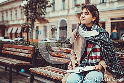 Cute shot of lovely girl sitting on a wooden bench outside, writing a poem, looking lost in thoughts and dreams. Stock Photo